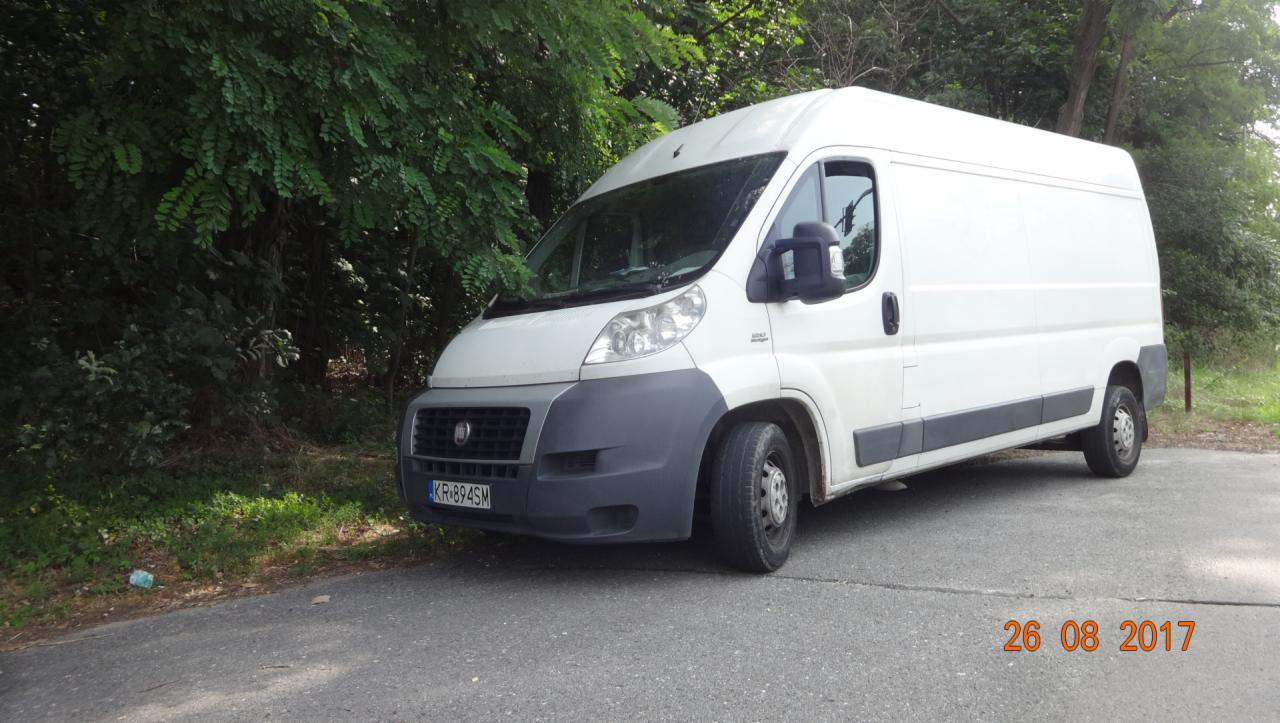 Biały samochód dostawczy marki Fiat Ducato zaparkowany na poboczu drogi asfaltowej, z widoczną tablicą rejestracyjną KR-894SM i datą 26 08 2017 w prawym dolnym rogu, otoczony zielenią drzew i krzewów.