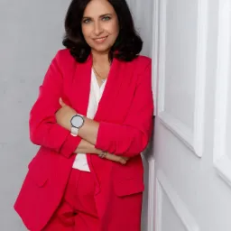 Portret uśmiechniętej kobiety w jaskrawoczerwonym garniturze, opierającej się o białą ścianę z dekoracyjnymi panelami, z zegarkiem na nadgarstku i delikatną biżuterią.