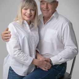 Atelier fotograficzne Duet Maciej Bazylewicz - Uśmiechnięta para w średnim wieku, kobieta z blond włosami i mężczyzna w kaszkiecie, oboje ubrani w białe koszule i dżinsy, pozują do portretu w studiu na tle jednolitego szarego tła.