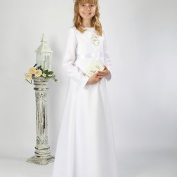 Atelier fotograficzne Duet Maciej Bazylewicz - Dziewczynka w białej albie komunijnej, z wiankiem na głowie, trzymająca książeczkę do Pierwszej Komunii Świętej, pozująca obok ozdobnej latarenki z kwiatami na białym tle.