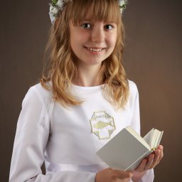 Atelier fotograficzne Duet Maciej Bazylewicz - Dziewczynka w białej albie komunijnej z wiankiem na głowie, trzymająca otwartą książkę na tle brązowego tła.
