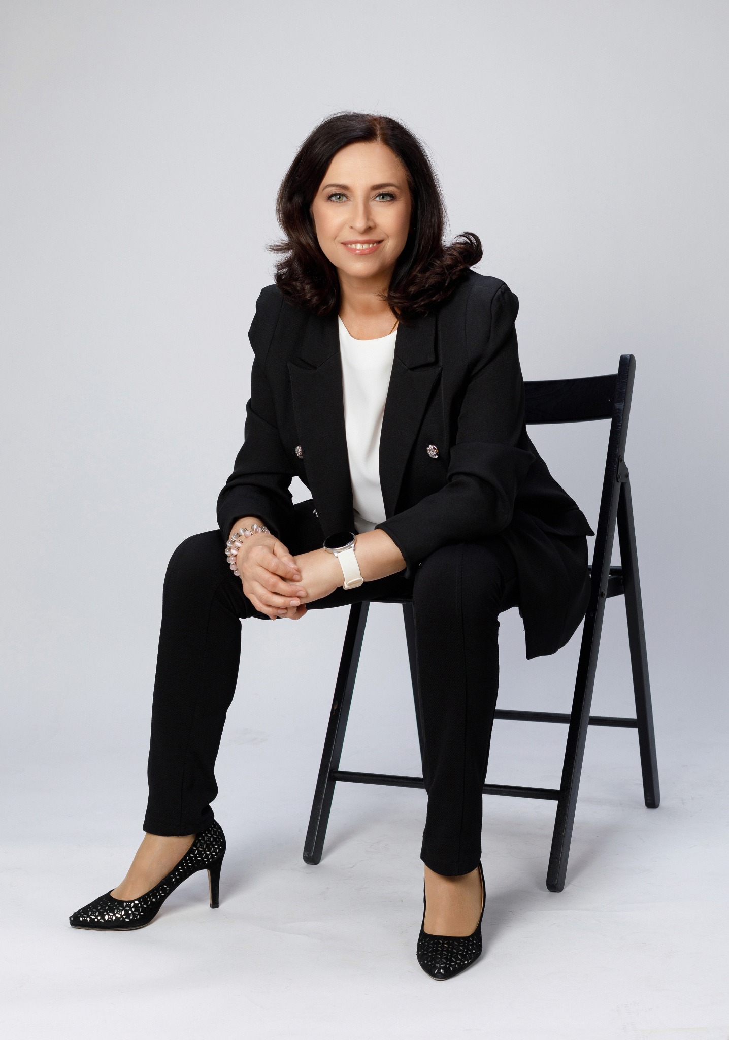 Kobieta w czarnym garniturze siedzi na krześle na jasnym tle, z delikatnym uśmiechem i elegancką biżuterią na nadgarstkach.