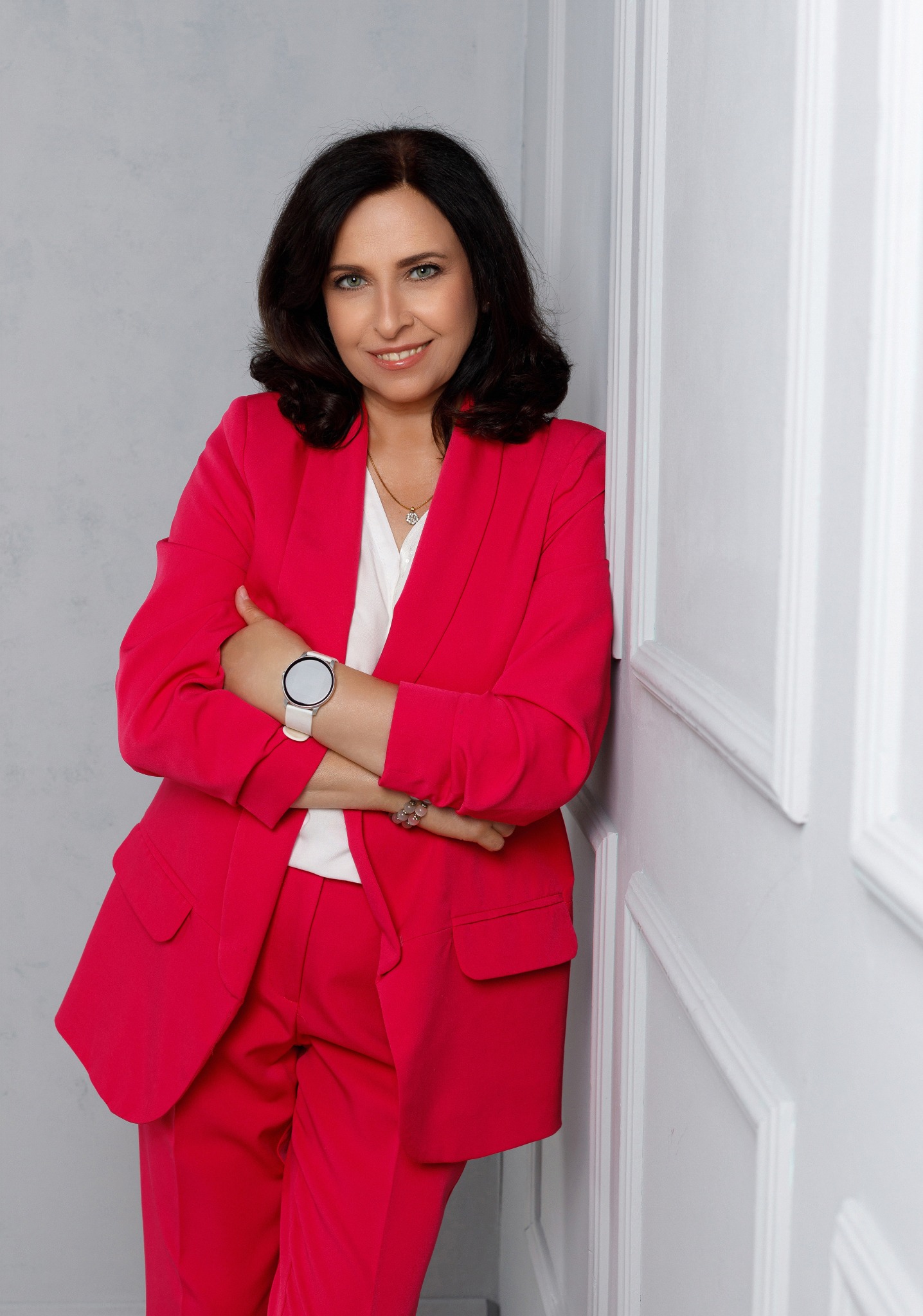Portret uśmiechniętej kobiety w jaskrawoczerwonym garniturze, opierającej się o białą ścianę z dekoracyjnymi panelami, z zegarkiem na nadgarstku i delikatną biżuterią.