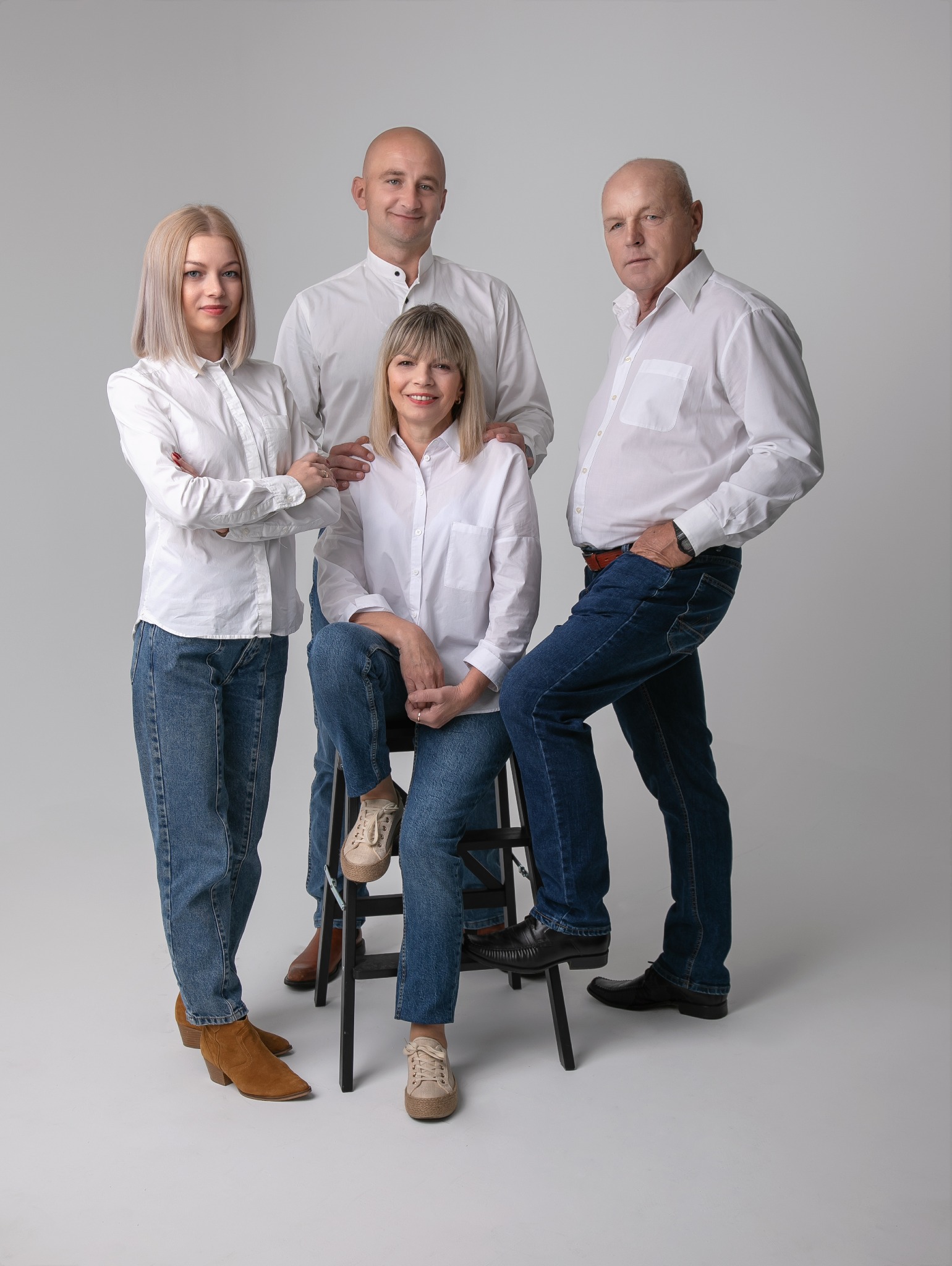Portret rodzinny: Dwie kobiety i dwóch mężczyzn w białych koszulach i jeansach pozują na jasnoszarym tle. Jedna z kobiet siedzi na czarnym stołku.