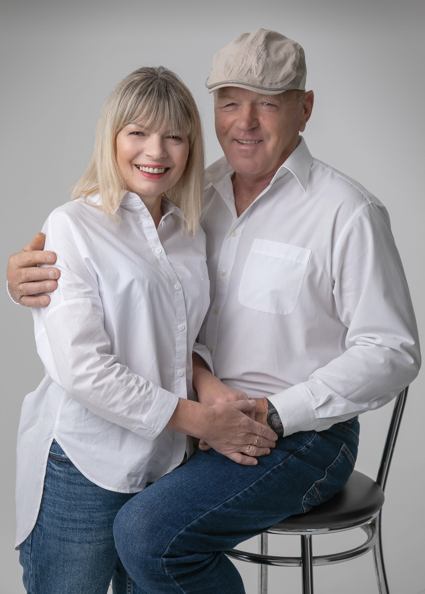 Uśmiechnięta para w średnim wieku, kobieta z blond włosami i mężczyzna w kaszkiecie, oboje ubrani w białe koszule i dżinsy, pozują do portretu w studiu na tle jednolitego szarego tła.