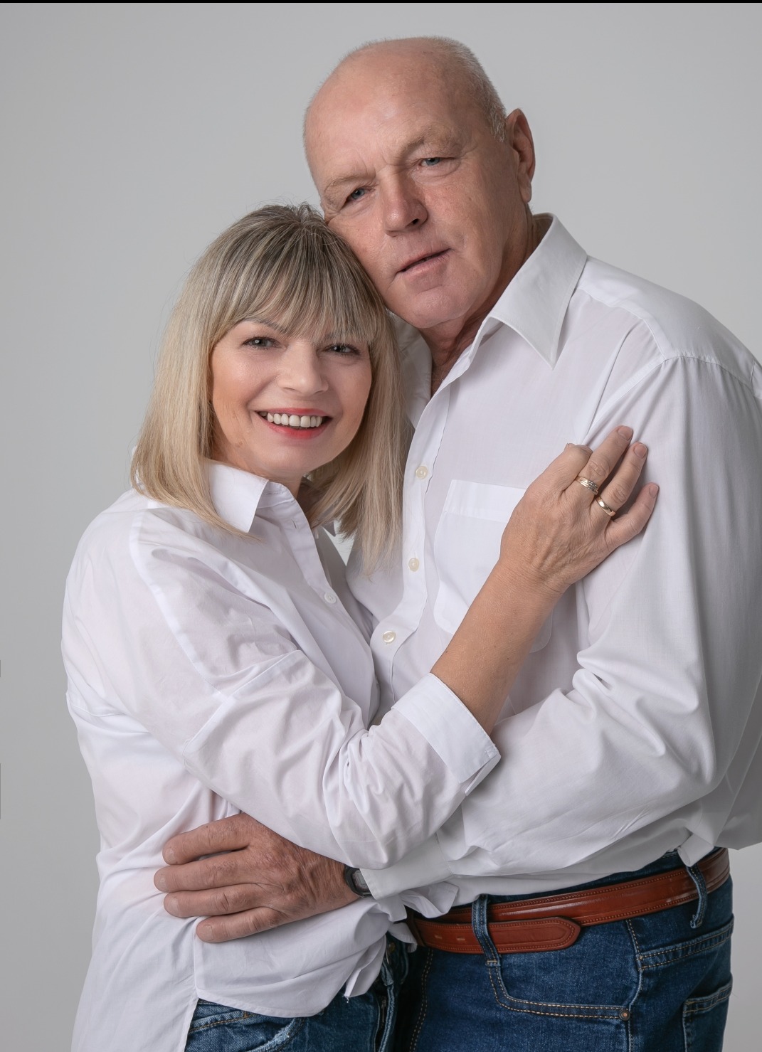 Uśmiechnięta para w średnim wieku, ubrana w białe koszule i jeansy, obejmująca się na jasnym tle. Widoczne obrączki ślubne.