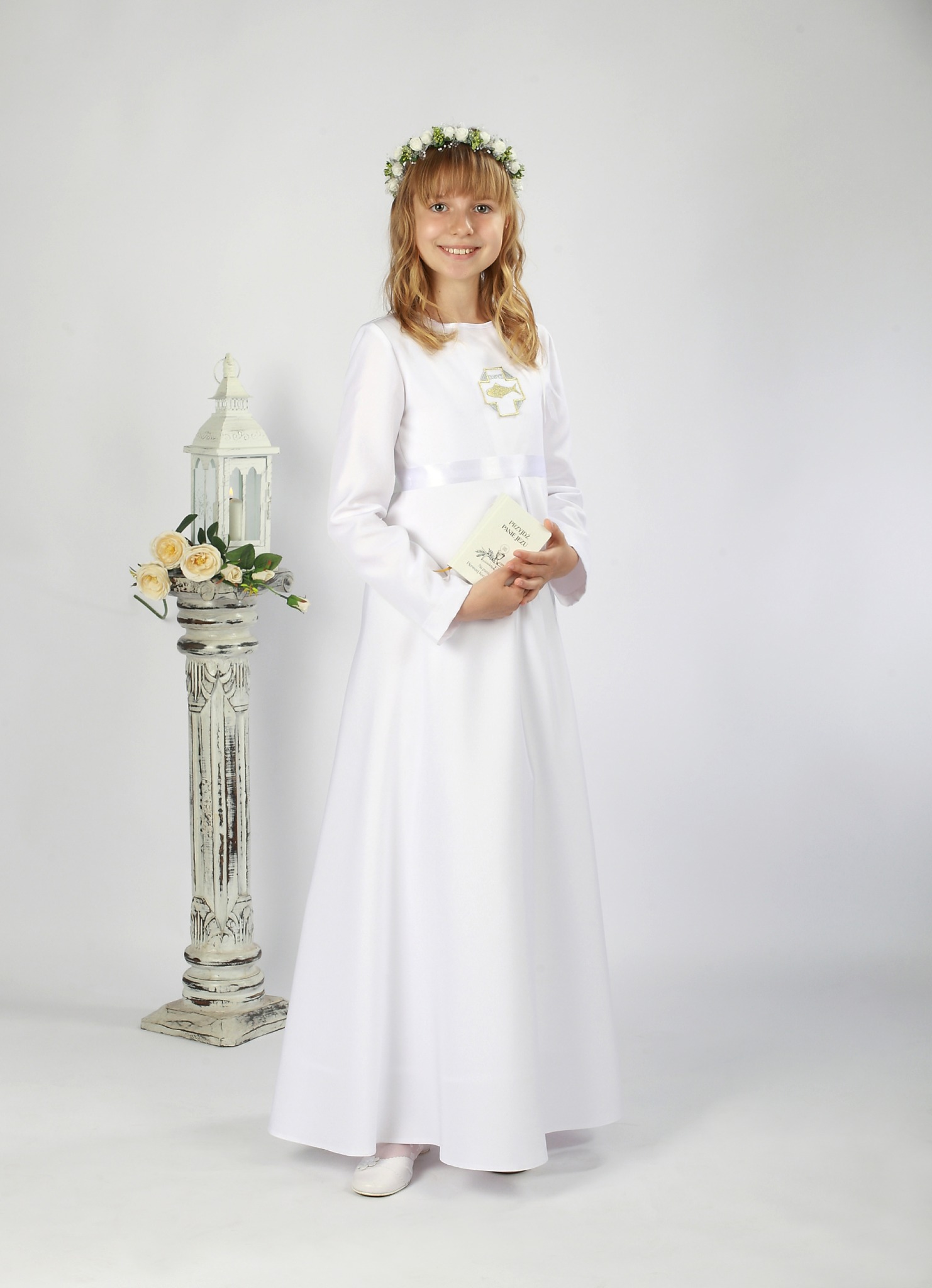 Dziewczynka w białej albie komunijnej, z wiankiem na głowie, trzymająca książeczkę do Pierwszej Komunii Świętej, pozująca obok ozdobnej latarenki z kwiatami na białym tle.