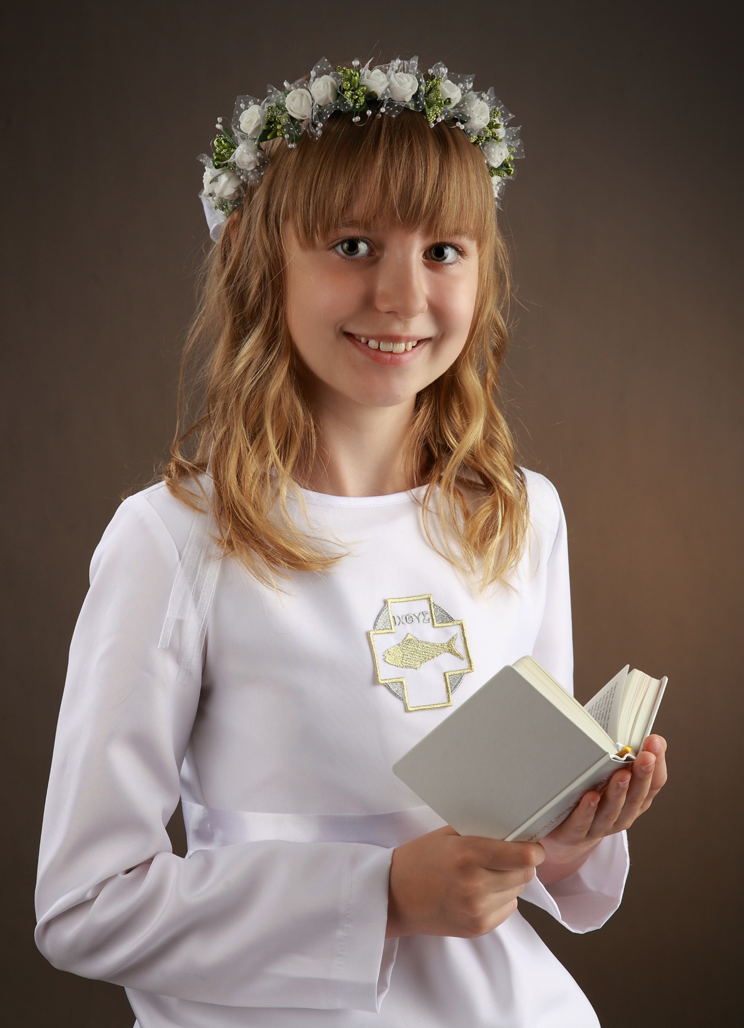 Dziewczynka w białej albie komunijnej z wiankiem na głowie, trzymająca otwartą książkę na tle brązowego tła.