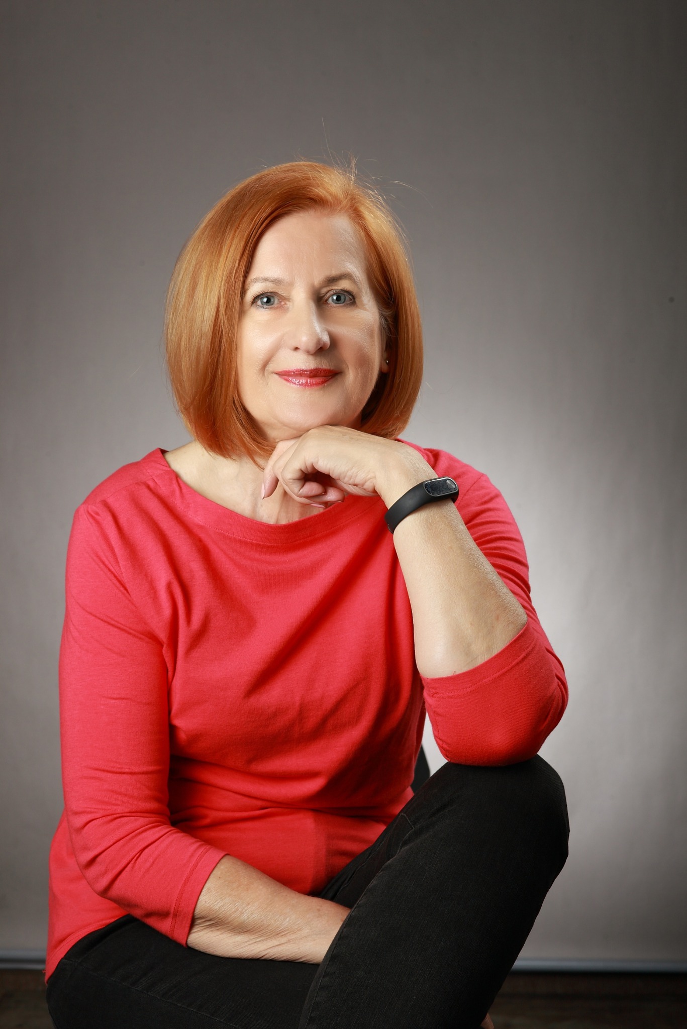 Portret uśmiechniętej kobiety z rudymi włosami, w czerwonej bluzce i czarnych spodniach, opierającej podbródek na dłoni z czarną opaską fitness, na szarym tle.