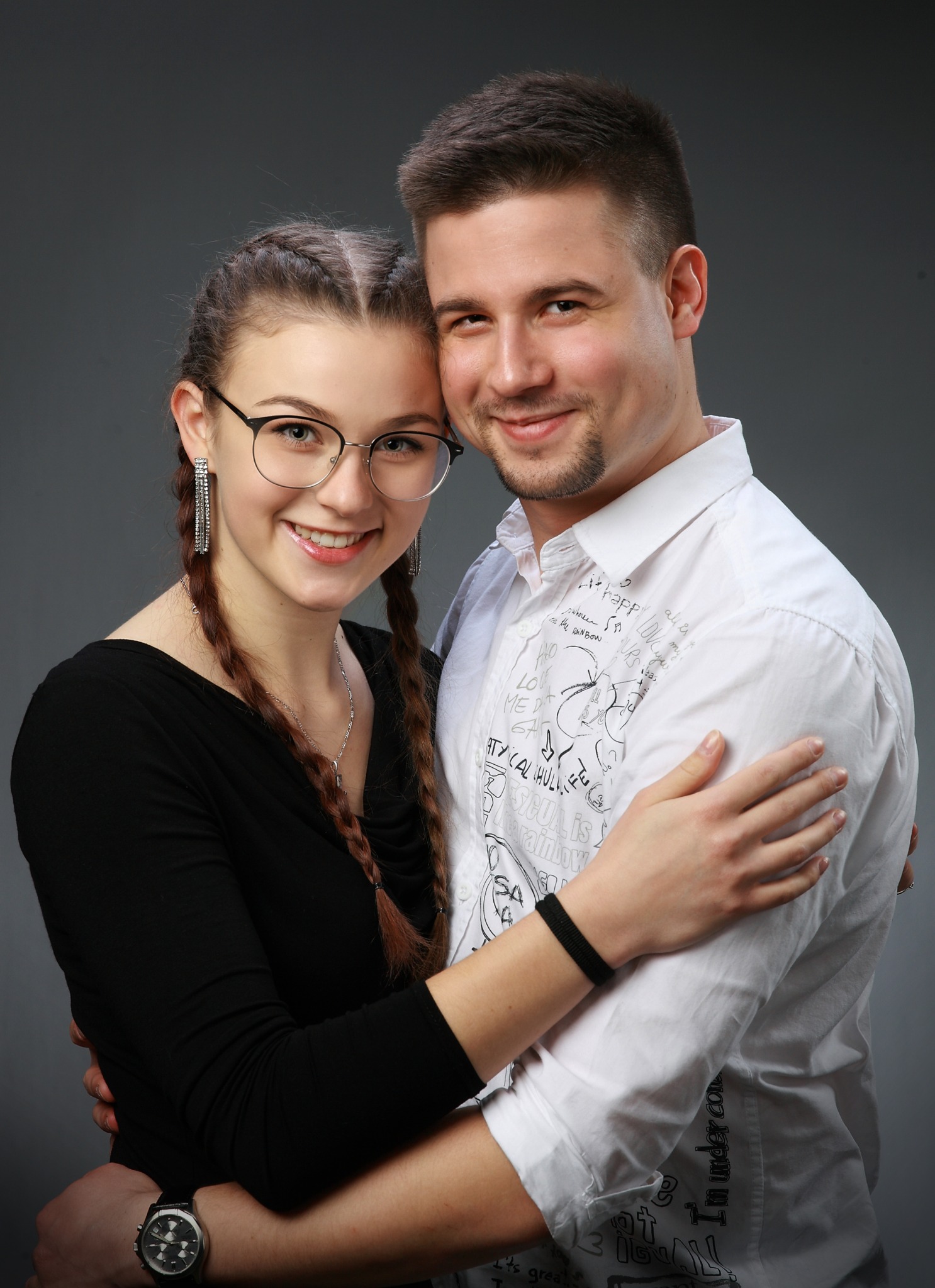 Uśmiechnięta para, dziewczyna w okularach i chłopak w koszuli z nadrukiem, obejmują się na szarym tle, widoczne detale biżuterii i zegarka.