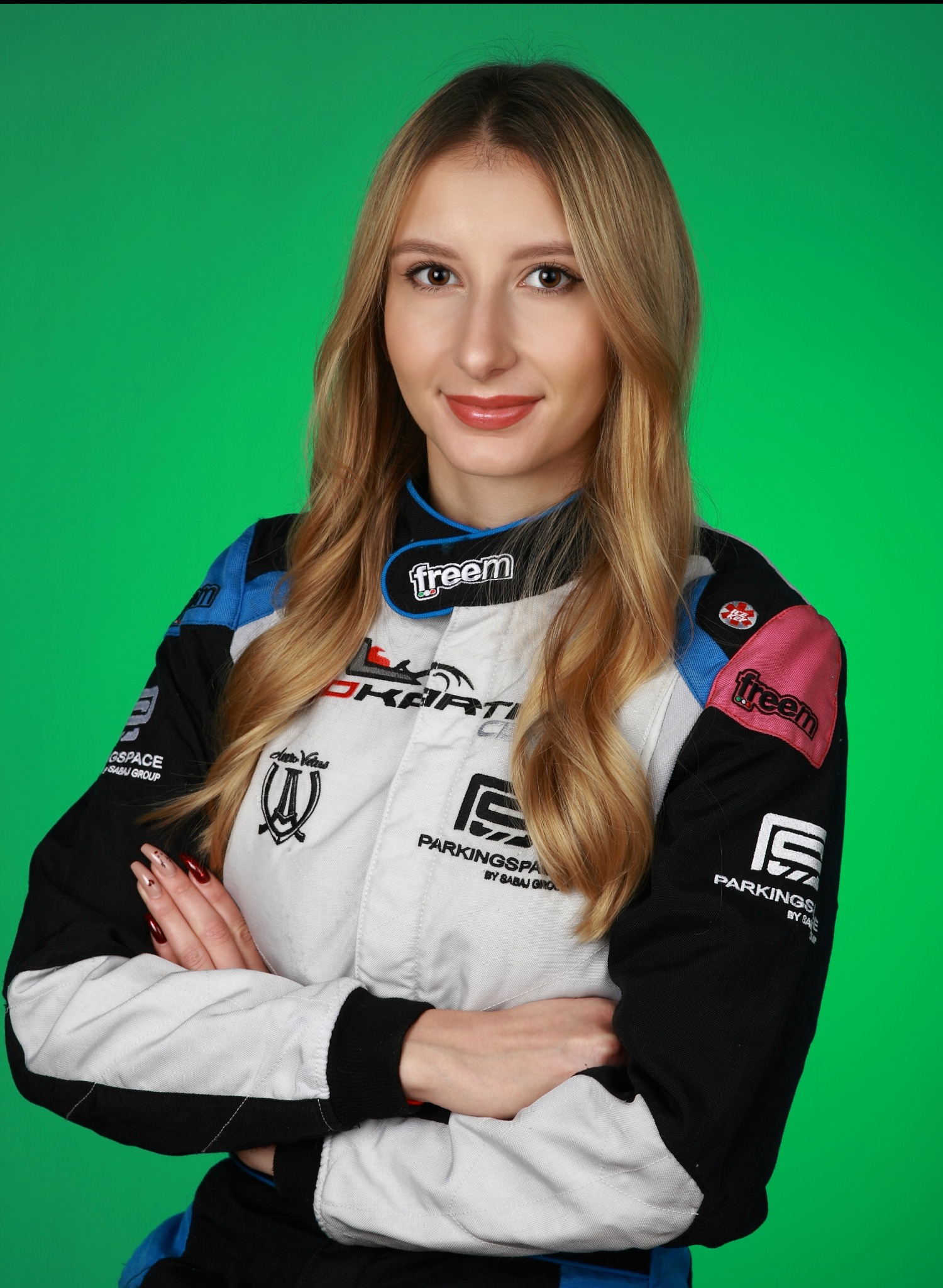 Portret uśmiechniętej kobiety w kombinezonie gokartowym z licznymi naszywkami sponsorskimi na tle zielonego ekranu.