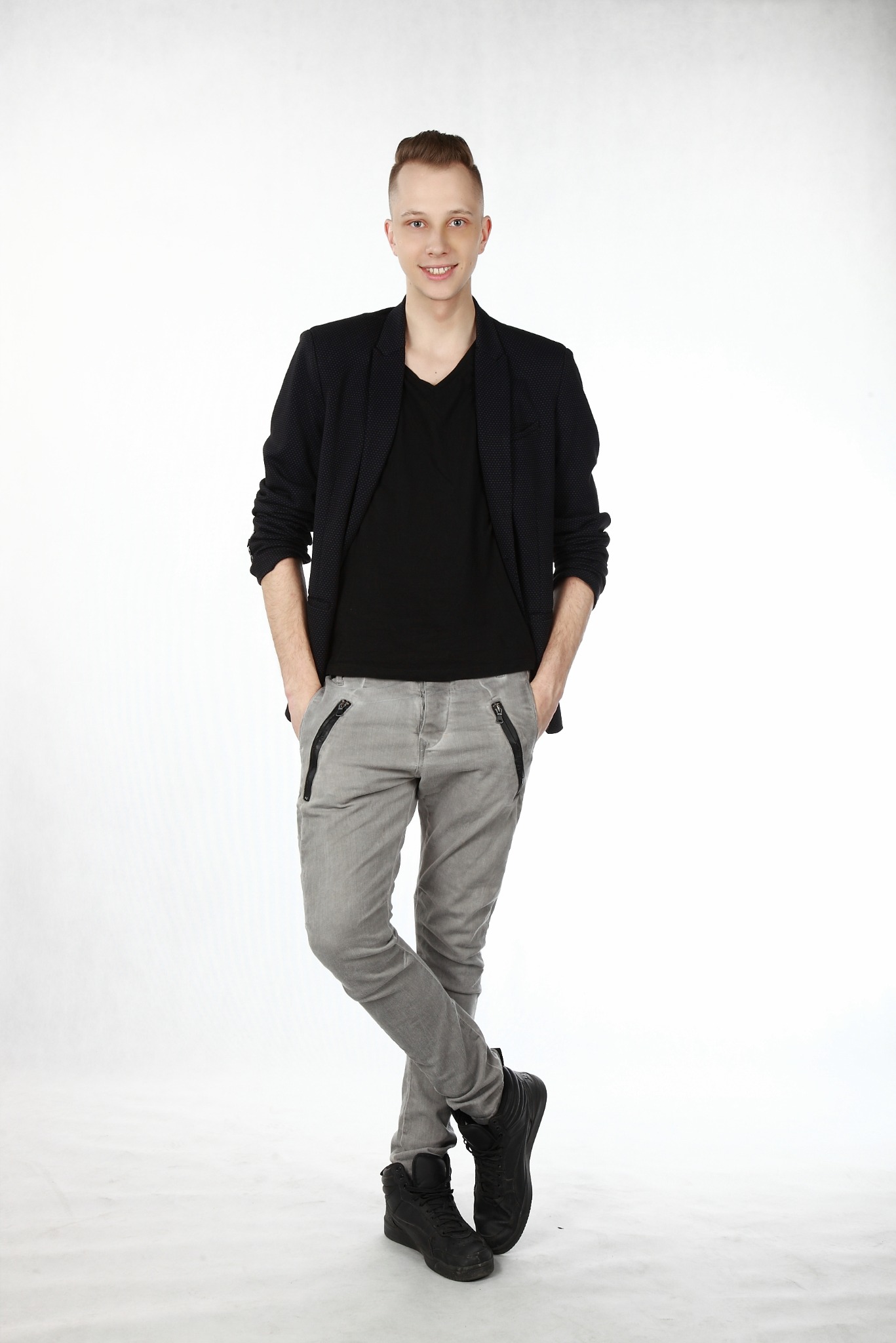 Młody mężczyzna w czarnej marynarce i szarych spodniach pozuje na białym tle w Krakowie, ręce w kieszeniach, nogi skrzyżowane.