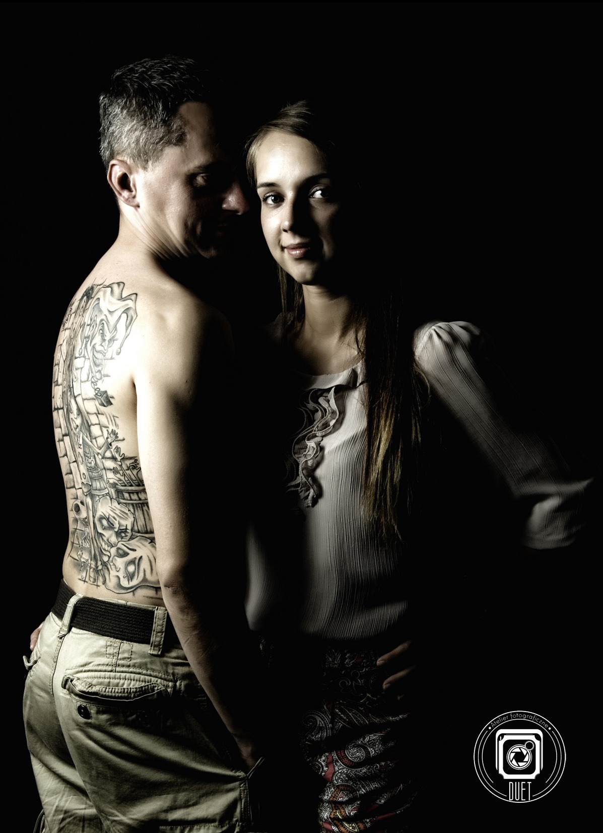 Portret pary w studiu z ciemnym tłem. Mężczyzna z tatuażem na plecach obejmuje kobietę. Oboje patrzą w obiektyw.