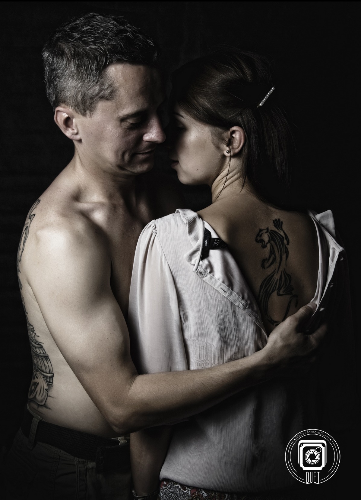 Intymne ujęcie pary w zbliżeniu na ciemnym tle; mężczyzna bez koszuli z tatuażem na plecach obejmuje kobietę z tatuażem pantery na odsłoniętych plecach.