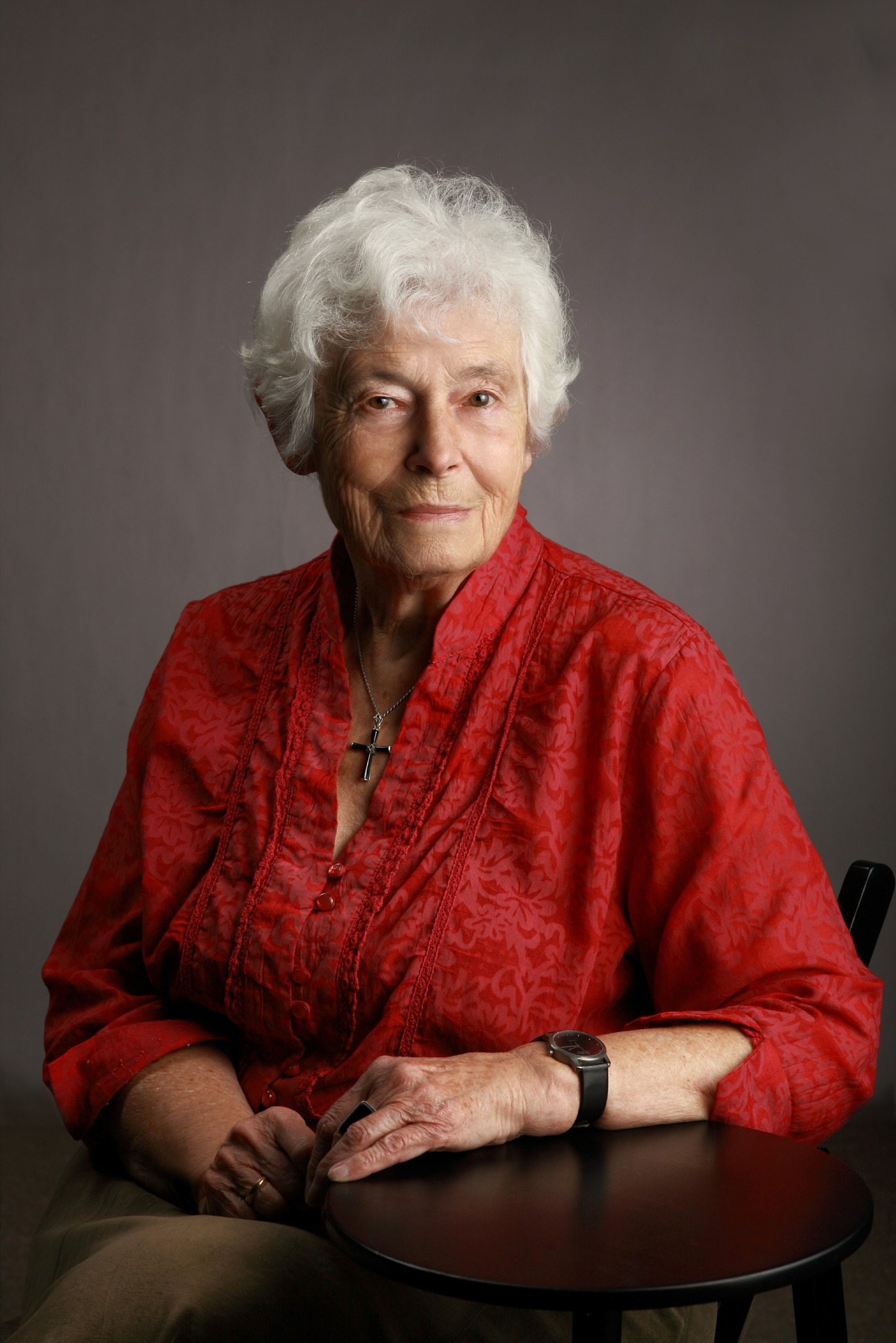 Portret starszej kobiety z siwymi włosami w czerwonej kwiecistej bluzce, siedzącej przy czarnym stoliku, z krzyżykiem na szyi i zegarkiem na nadgarstku, na szarym tle.