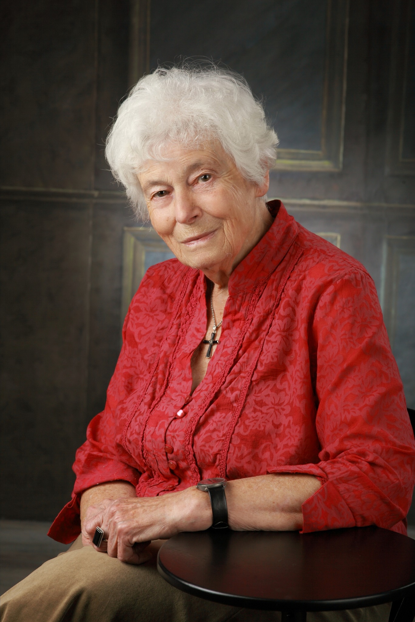 Portret uśmiechniętej starszej kobiety w czerwonej bluzce z wzorem, siedzącej na krześle z czarnym zegarkiem i pierścionkiem na palcu, na tle abstrakcyjnej ściany.