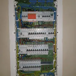Otwarta skrzynka elektryczna z zainstalowanymi bezpiecznikami, różnicówkami i przewodami o różnych kolorach, zamontowana na ścianie wewnątrz budynku.