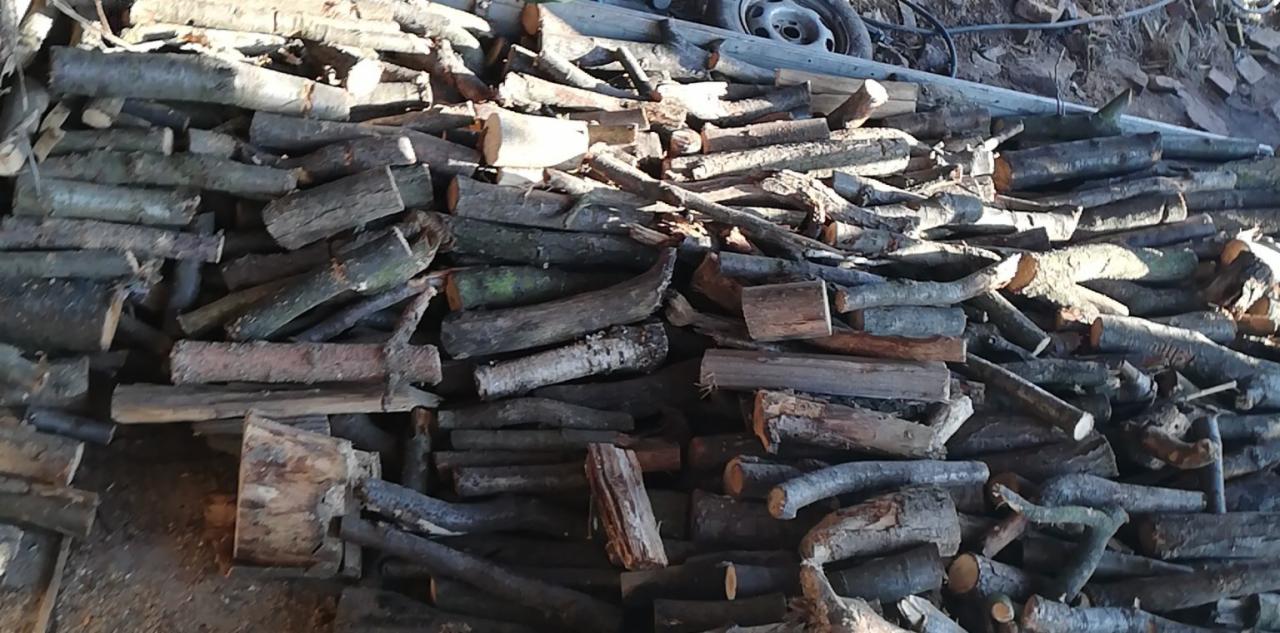 Stos pociętego drewna opałowego w nieładzie, widoczne fragmenty opon samochodowych w tle.