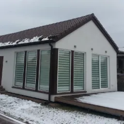 Nowoczesny, parterowy dom z białymi ścianami i brązowymi ramami okiennymi, częściowo pokryty śniegiem, z widocznym tarasem i schodami wejściowymi.