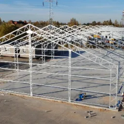 Konstrukcja stalowa hali w trakcie budowy, widoczne elementy szkieletu, zadaszenie w budowie, na zewnątrz budynki gospodarcze i drzewa.