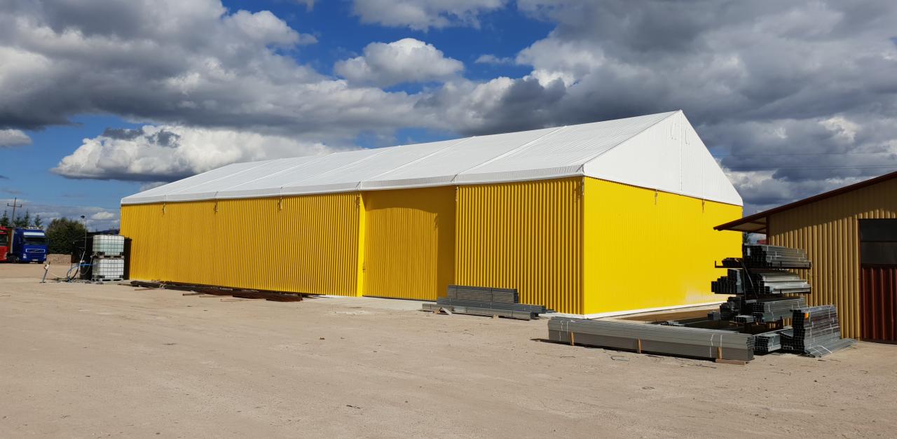 Żółta hala magazynowa z białym dachem, widok z zewnątrz w słoneczny dzień, z elementami konstrukcyjnymi składowanymi obok.