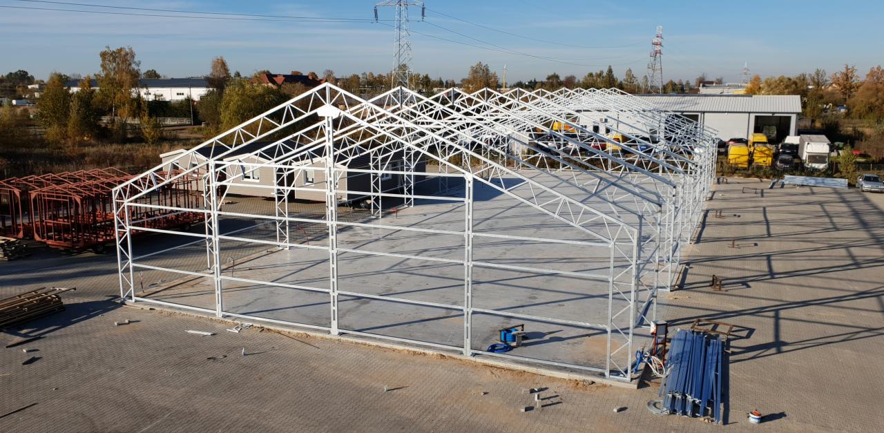 Konstrukcja stalowa hali w trakcie budowy, widoczne elementy szkieletu, zadaszenie w budowie, na zewnątrz budynki gospodarcze i drzewa.