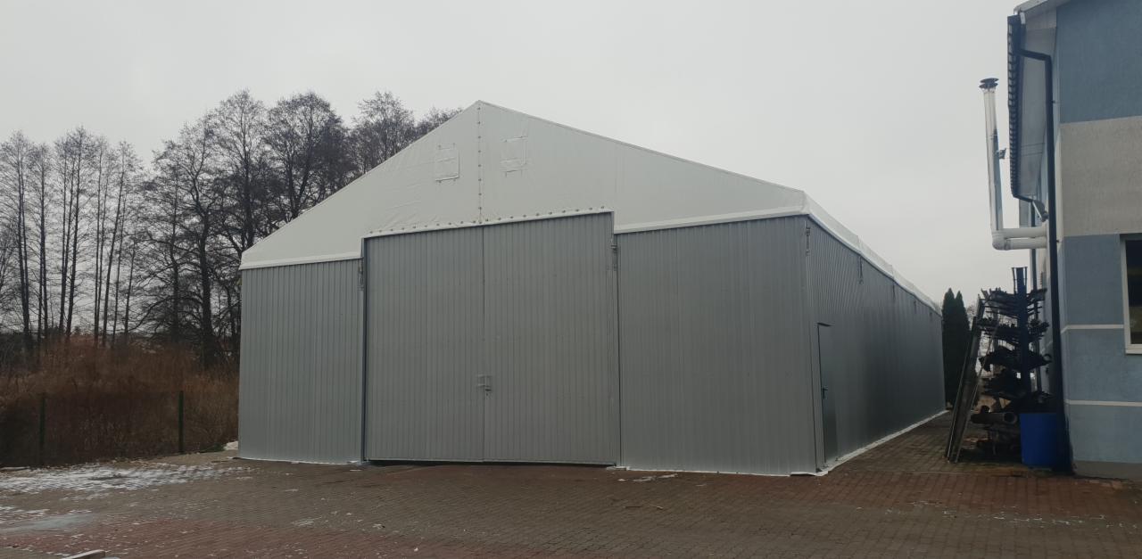 Gotowa hala magazynowa z szarej blachy trapezowej i białym dachem, widoczne duże przesuwne wrota i małe drzwi wejściowe, obok budynek z kominem i stertą rur.