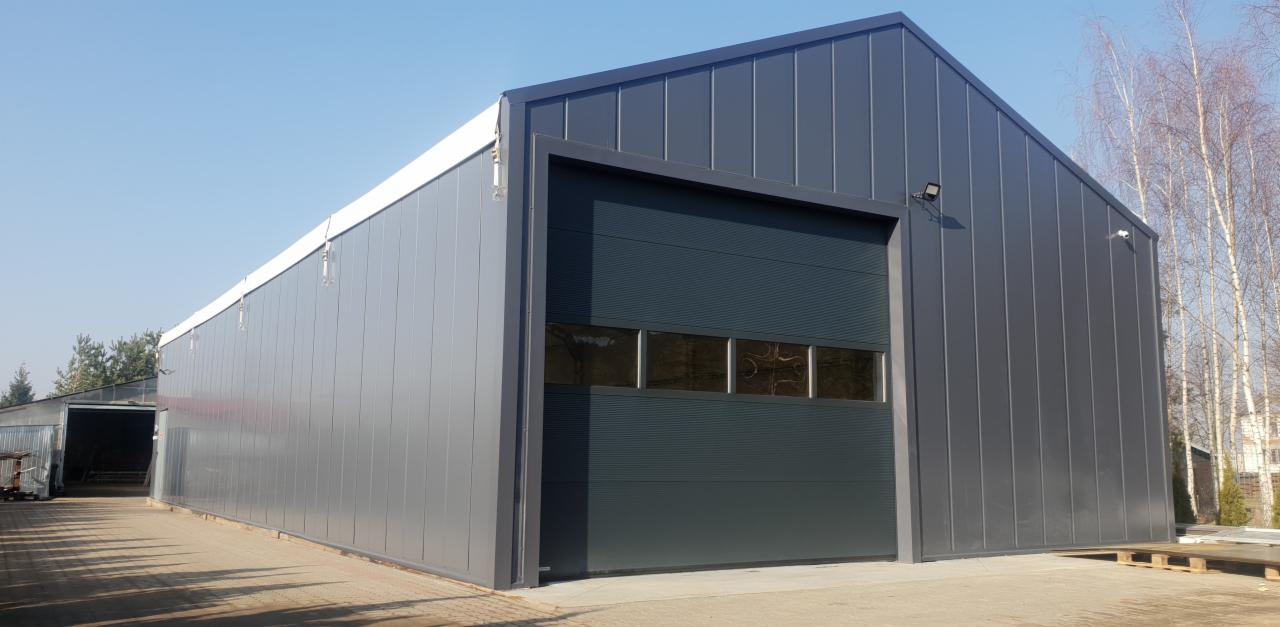 Zewnętrzny widok szarej hali stalowej z segmentową bramą garażową z oknami, oświetleniem zewnętrznym i widocznymi drzewami w tle.