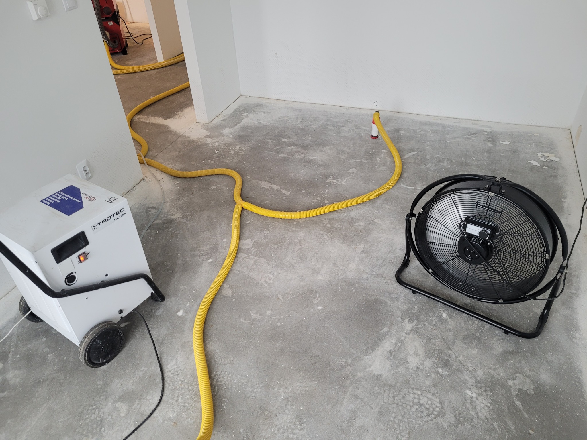 Pokój w trakcie osuszania: biały osuszacz powietrza Trotec, żółty wąż odprowadzający wilgoć i czarny wentylator stojący na posadzce z widocznymi śladami wilgoci na ścianach.