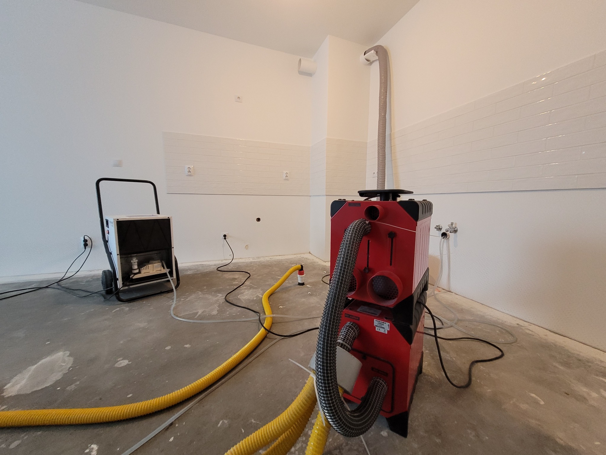 Dwa urządzenia do osuszania w pomieszczeniu z nowymi płytkami na ścianie i surową, betonową podłogą. Jeden osuszacz czerwony z grubym, elastycznym przewodem, drugi biały na kółkach.