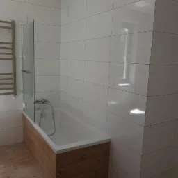 Biała łazienka z wanną obudowaną drewnopodobnymi panelami, szklanym parawanem nawannowym i grzejnikiem drabinkowym na ścianie z białych płytek, drewniana podłoga.