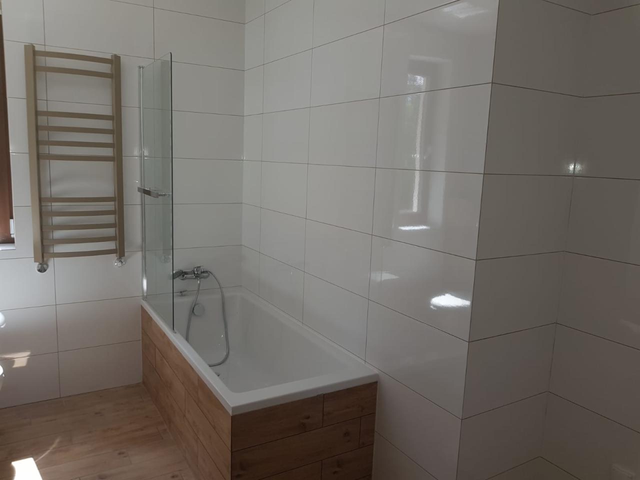 Biała łazienka z wanną obudowaną drewnopodobnymi panelami, szklanym parawanem nawannowym i grzejnikiem drabinkowym na ścianie z białych płytek, drewniana podłoga.