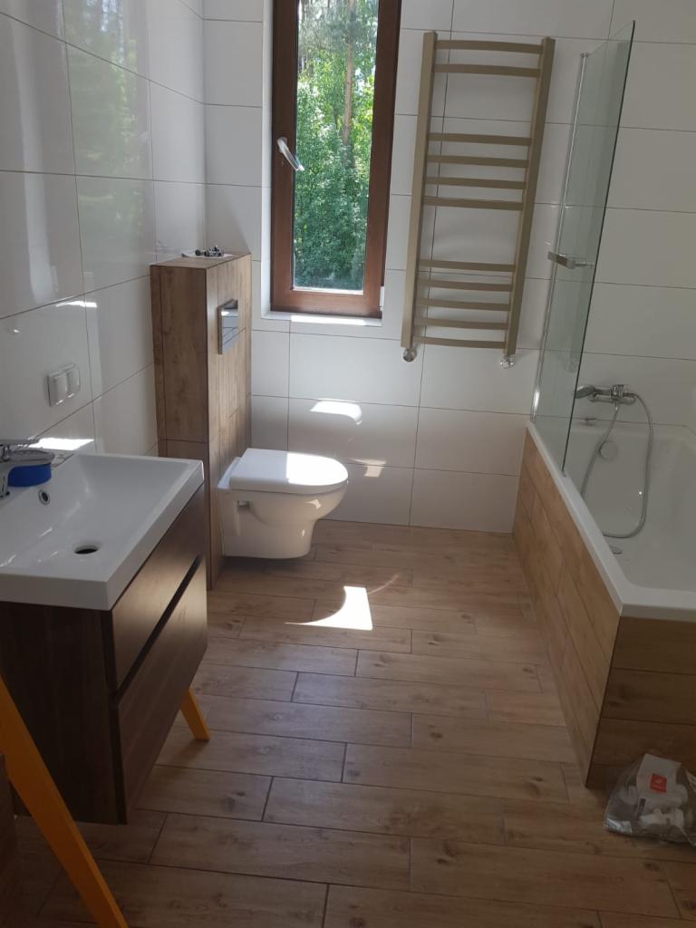 Jasna łazienka z drewnianymi akcentami: podłoga, obudowa wanny i szafka pod umywalką, grzejnik drabinkowy, okno z widokiem na zieleń.