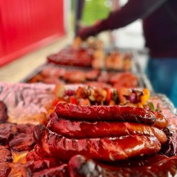 ADAM STRYCHARZ - Grillowane kiełbaski ułożone na folii aluminiowej, w tle szaszłyki i inne mięsa na ruszcie, przygotowywane przez osobę w ciemnej bluzie.