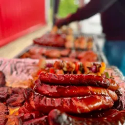 Grillowane kiełbaski ułożone na folii aluminiowej, w tle szaszłyki i inne mięsa na ruszcie, przygotowywane przez osobę w ciemnej bluzie.