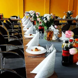 Długi stół nakryty szarym obrusem, zastawiony białymi talerzami z potrawami, szklankami, napojami w butelkach i wazonami z tulipanami, ustawiony w sali z żółtymi ścianami.
