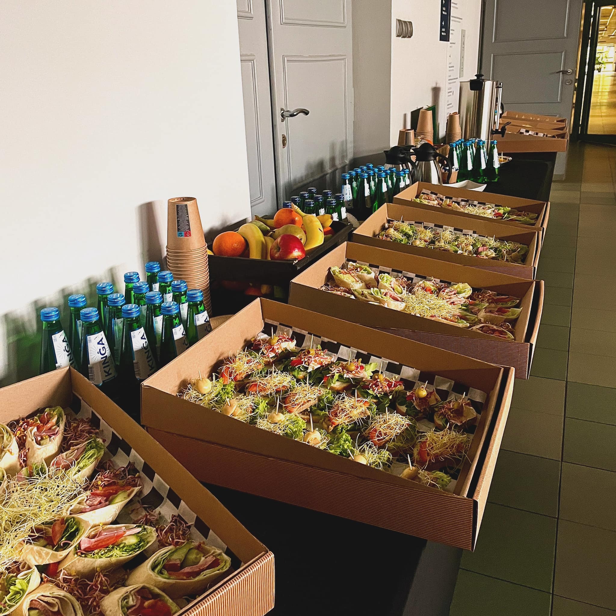 Długi stół z przekąskami w kartonowych pudełkach z wzorem szachownicy, butelkami wody Kinga i owocami, ustawiony w jasnym pomieszczeniu z szarymi drzwiami.