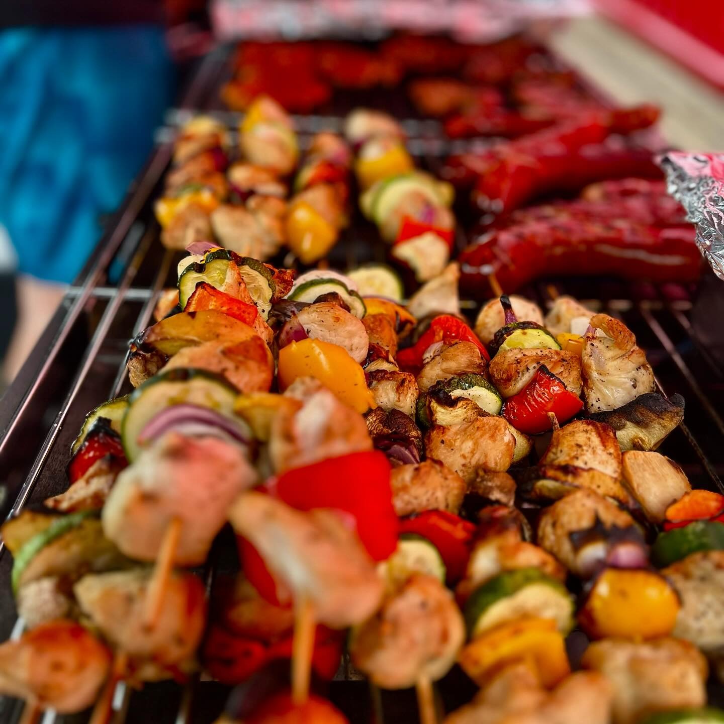 Grillowane szaszłyki z kurczaka i warzyw (papryka, cukinia, cebula) na ruszcie, z widocznymi śladami grillowania, w tle kolejne szaszłyki i czerwone kiełbaski.