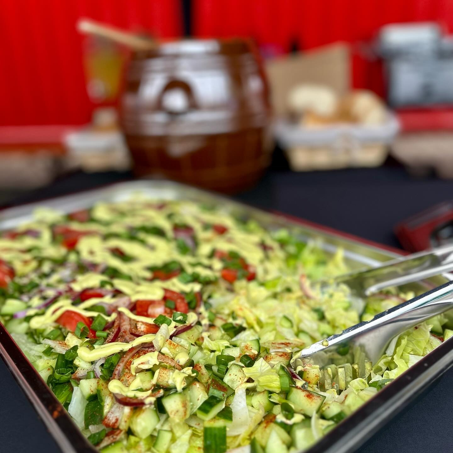 Duża metalowa taca pełna świeżej sałatki z ogórkiem, pomidorem, czerwoną cebulą i sosem, szczypce do nakładania, w tle gliniany garnek i koszyk z pieczywem.