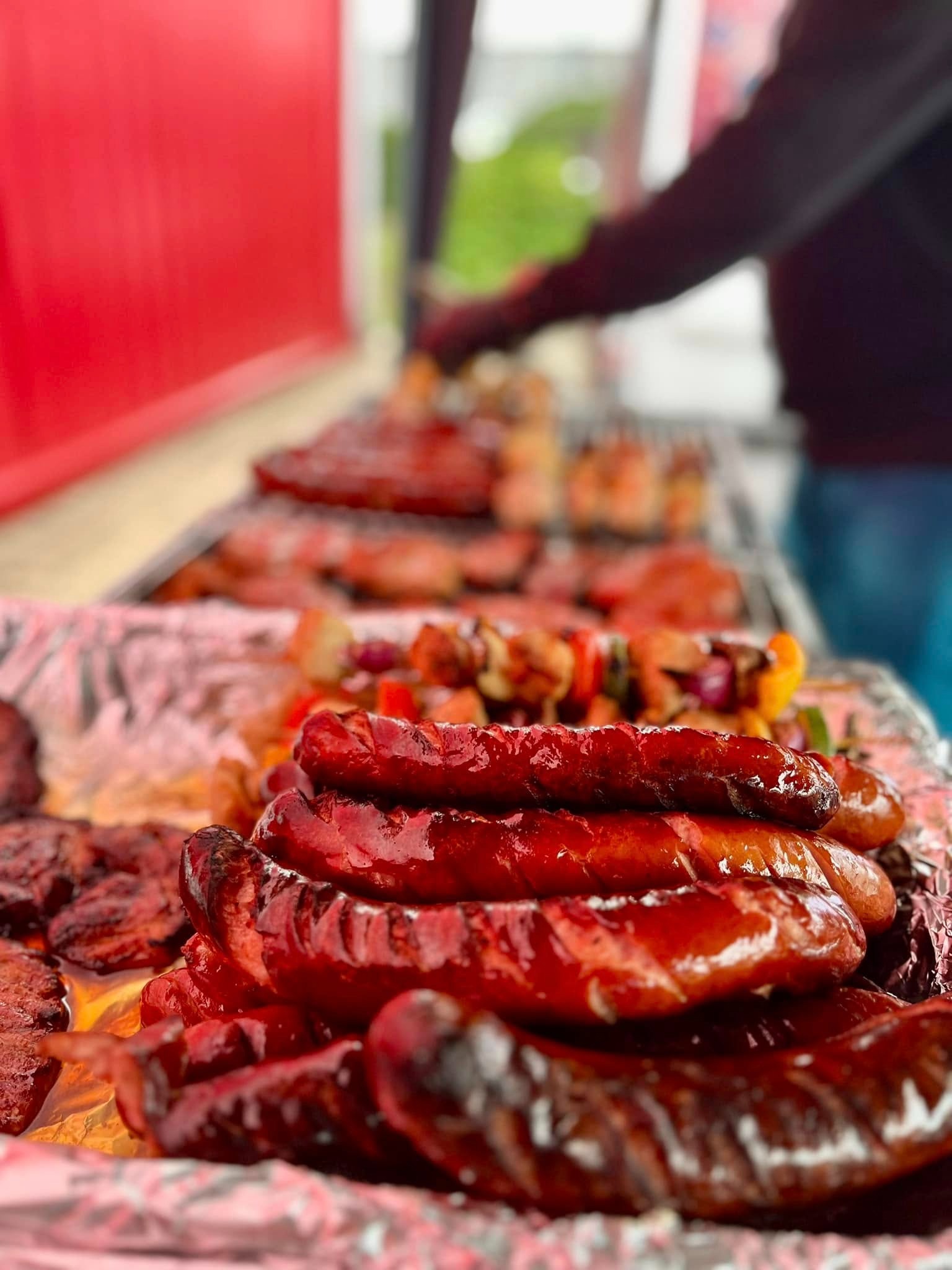 Grillowane kiełbaski ułożone na folii aluminiowej, w tle szaszłyki i inne mięsa na ruszcie, przygotowywane przez osobę w ciemnej bluzie.