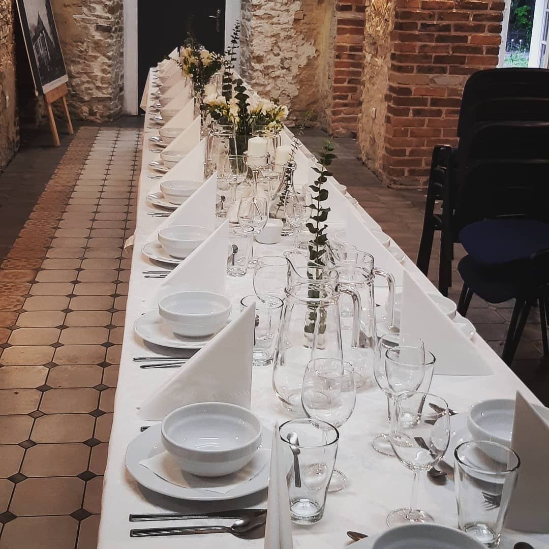 Długi stół nakryty białym obrusem, z białymi naczyniami, szklankami i dzbankami z gałązkami eukaliptusa, ustawiony w sali z ceglanymi ścianami i płytkami na podłodze, przygotowany na uroczystość.