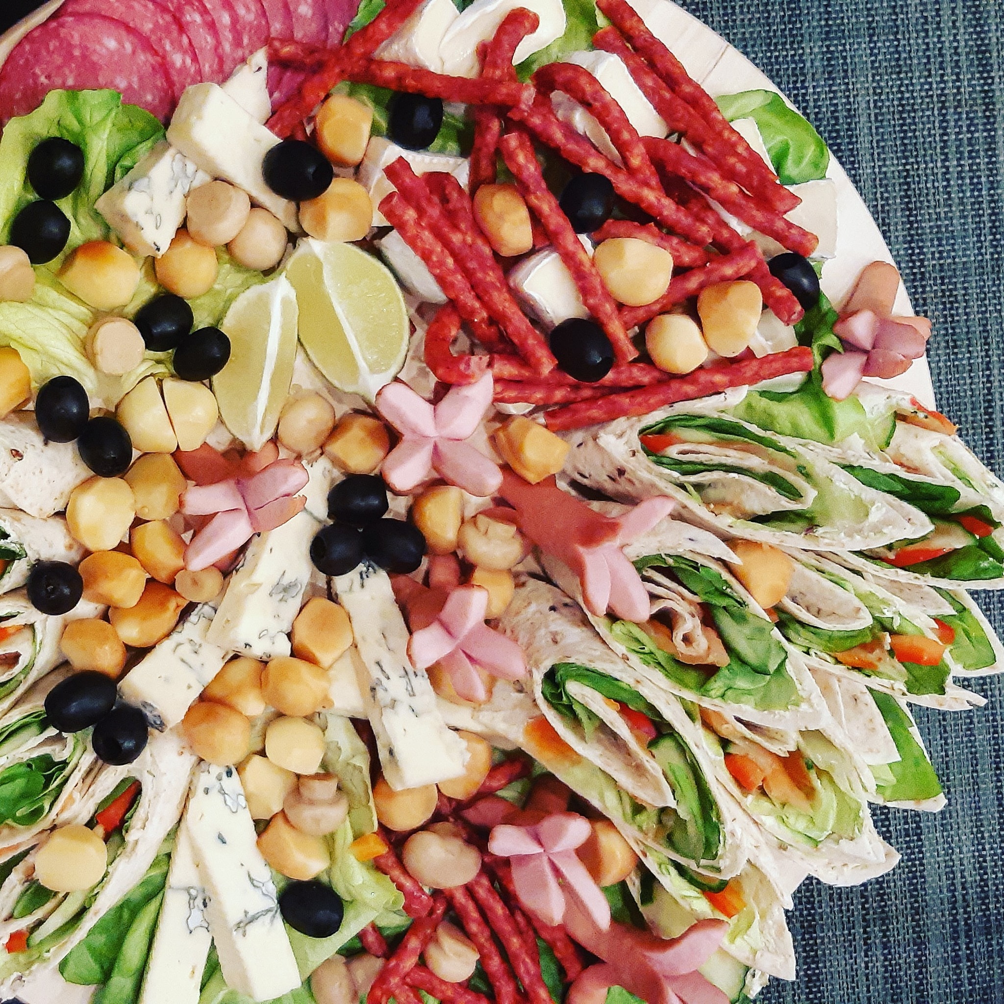 Dekoracyjna taca z przekąskami: sery pleśniowe, salami, kabanosy, macadamia, oliwki, limonka, wrap z warzywami i dekoracje z wędliny w kształcie kwiatów.