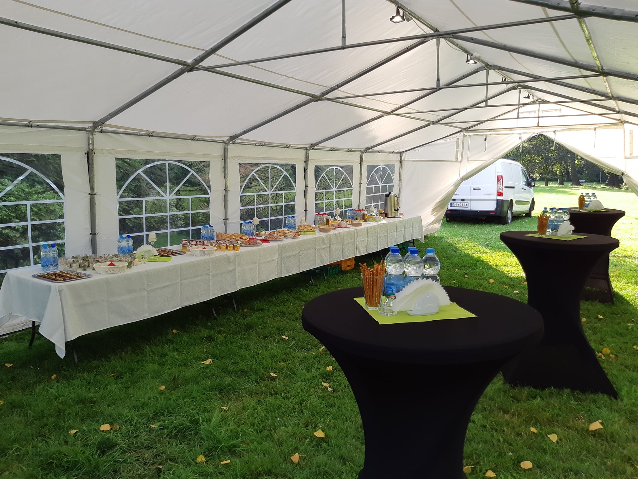 Uroczysty bufet z przekąskami i napojami ustawiony wewnątrz białego namiotu plenerowego, z widocznymi stolikami koktajlowymi, catering na trawie, biały samochód dostawczy w tle.