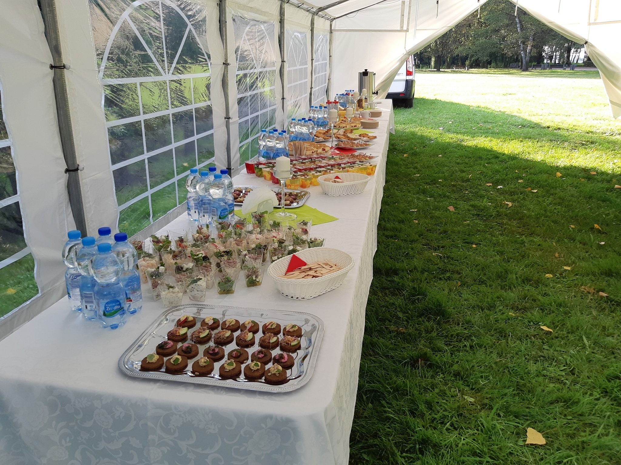 Stół cateringowy pod namiotem, z wodą butelkowaną, małymi przekąskami w szklanych naczyniach, kanapeczkami i ciastkami na srebrnej tacy, ustawiony na białym obrusie w plenerze.