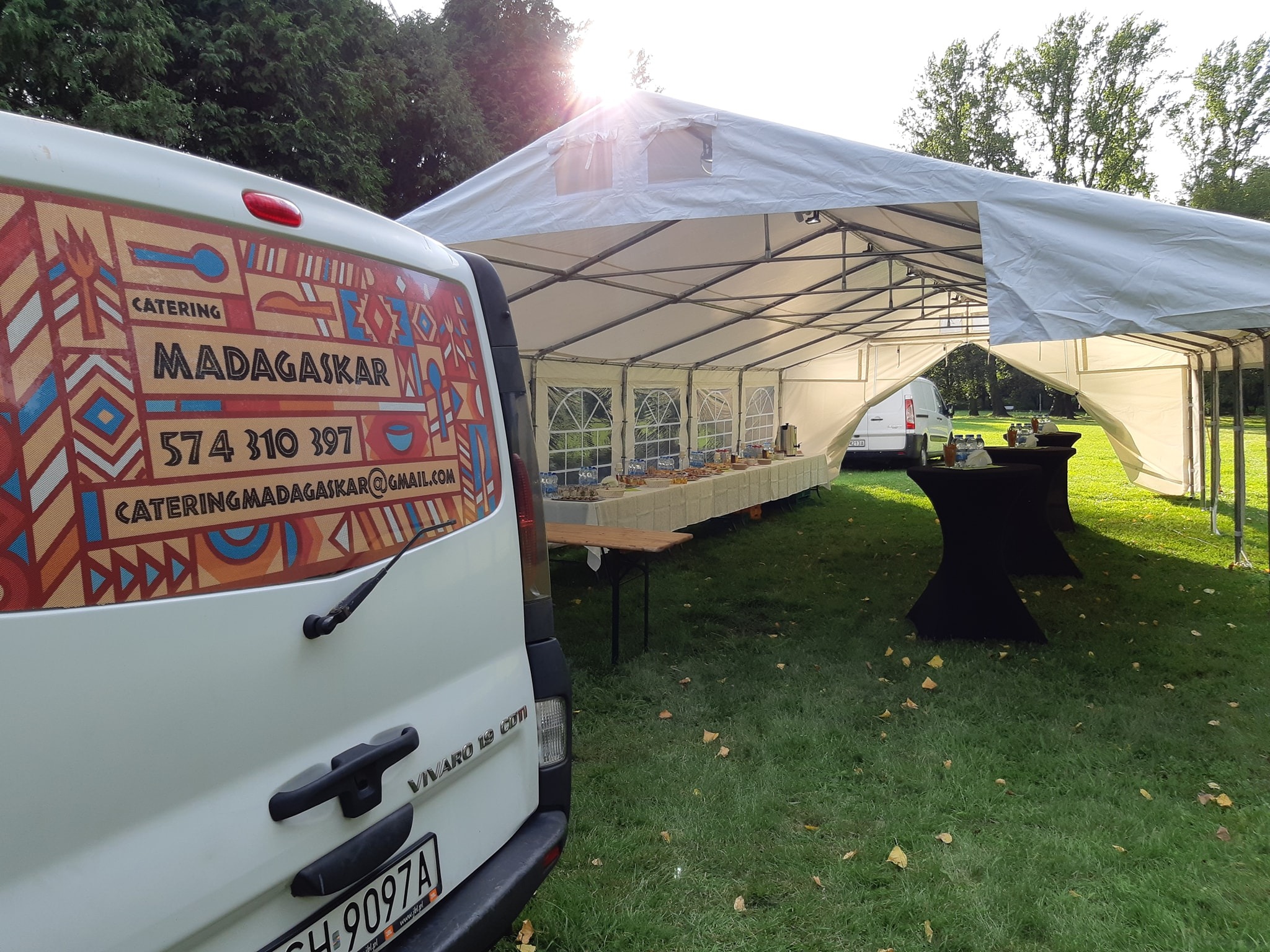 Biały namiot cateringowy z bufetem widoczny zza furgonetki z kolorową grafiką i danymi kontaktowymi firmy cateringowej Madagaskar, na zielonym trawniku.