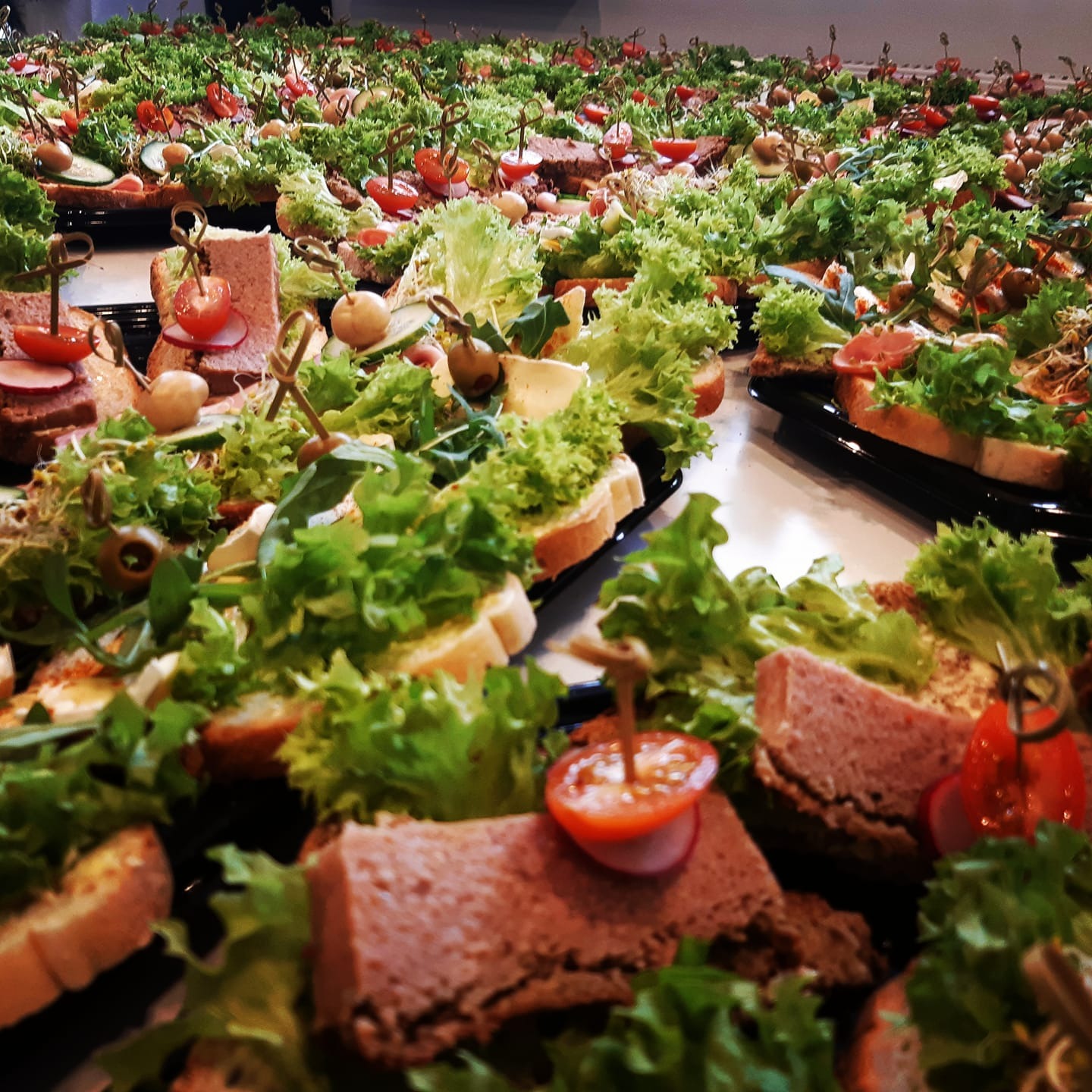 Rozległy widok na liczne, apetycznie wyglądające kanapeczki na ciemnych tacach, udekorowane świeżą sałatą, pomidorkami cherry i oliwkami, niektóre z pastą, inne z wędlinami, wszystkie z ozdobnymi...