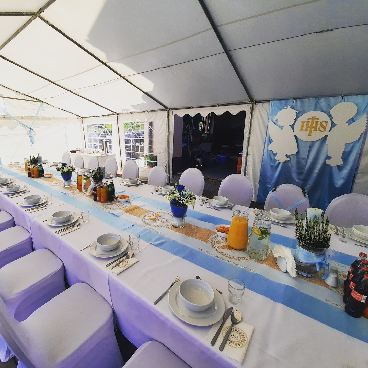 Uroczysty stół komunijny pod namiotem: biały obrus z niebieskim bieżnikiem, dekoracje kwiatowe w niebieskich wazonach, napoje w szklanych dzbankach i butelkach, biała zastawa, serwetki z symbolem...