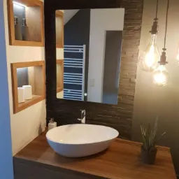 Nowoczesna łazienka z owalną białą umywalką na drewnianym blacie, czarną ścianą z kamienia dekoracyjnego, podświetlanymi półkami wnękowymi i wiszącymi żarówkami dekoracyjnymi.