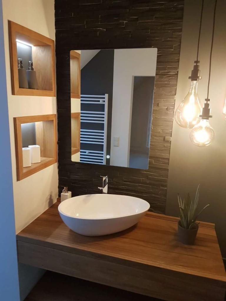 Nowoczesna łazienka z owalną białą umywalką na drewnianym blacie, czarną ścianą z kamienia dekoracyjnego, podświetlanymi półkami wnękowymi i wiszącymi żarówkami dekoracyjnymi.