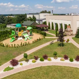 Widok z lotu ptaka na plac zabaw z zielonymi zjeżdżalniami i piaskownicą, część terenu rekreacyjnego z pawilonami i ścieżkami spacerowymi przy Sali Bankietowej w słoneczny dzień.